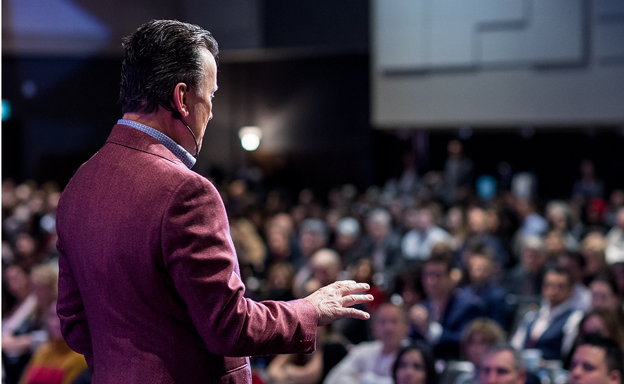 Man in red jacket speaking to a large audience during a conference