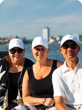 Three adults in white caps smiling together outdoors with a waterfront cityscape behind them.